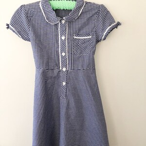 Puede incluir: Un vestido de cuadros azules y blancos con cuello Peter Pan, mangas cortas y abotonadura frontal. El vestido tiene ribetes blancos en el cuello, bolsillo y tapeta de botones. Cuelga de una percha blanca.