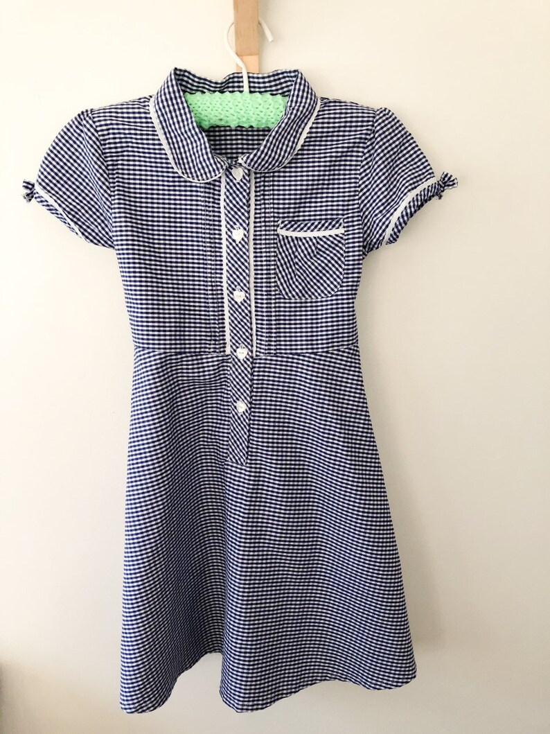 Puede incluir: Un vestido de cuadros azules y blancos con cuello Peter Pan, mangas cortas y abotonadura frontal. El vestido tiene ribetes blancos en el cuello, bolsillo y tapeta de botones. Cuelga de una percha blanca.