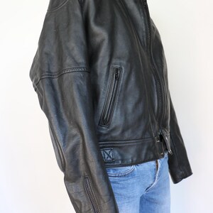 May include: Black leather jacket with a stand-up collar, zipper closure, and zippered pockets. The jacket has a classic biker style with a fitted silhouette. The sleeves have zippers at the cuffs. The jacket is paired with blue jeans.