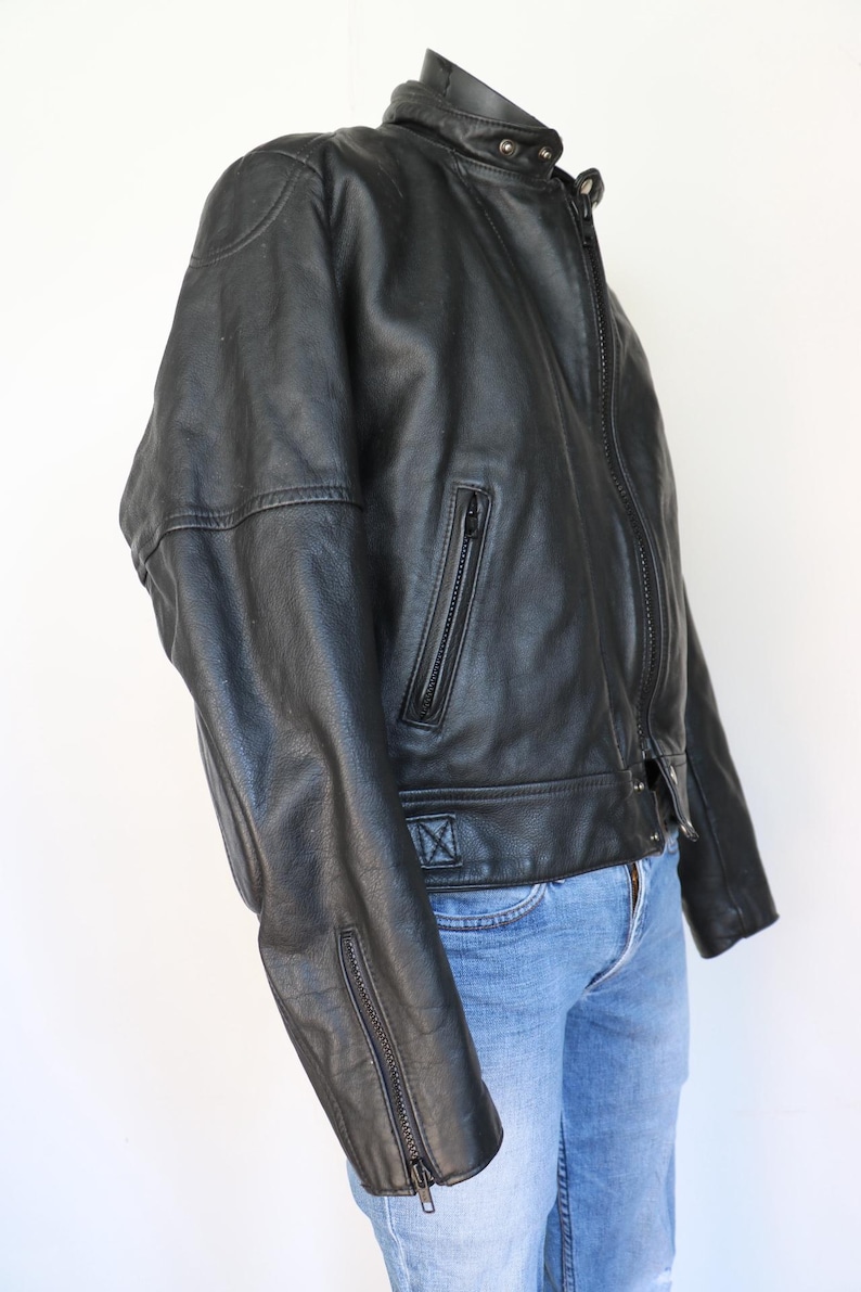 May include: Black leather jacket with a stand-up collar, zipper closure, and zippered pockets. The jacket has a classic biker style with a fitted silhouette. The sleeves have zippers at the cuffs. The jacket is paired with blue jeans.