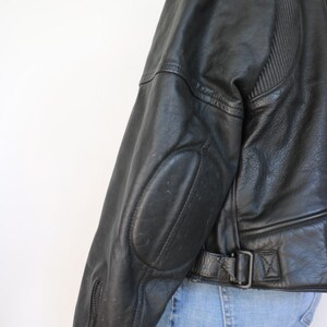 May include: Black leather jacket with ribbed shoulder detailing and elbow patches. Features a zippered sleeve and a belt with a buckle at the waist. The jacket is worn with blue jeans.