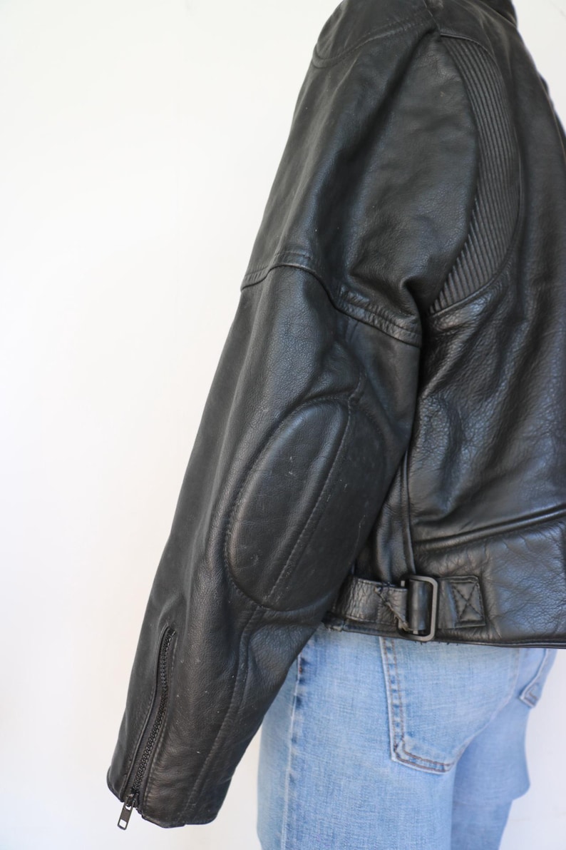 May include: Black leather jacket with ribbed shoulder detailing and elbow patches. Features a zippered sleeve and a belt with a buckle at the waist. The jacket is worn with blue jeans.