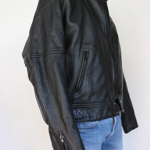May include: Black leather motorcycle jacket with a stand-up collar, zippered pockets, and sleeve zippers. The jacket has a classic design with a fitted silhouette. The jacket is paired with blue jeans.