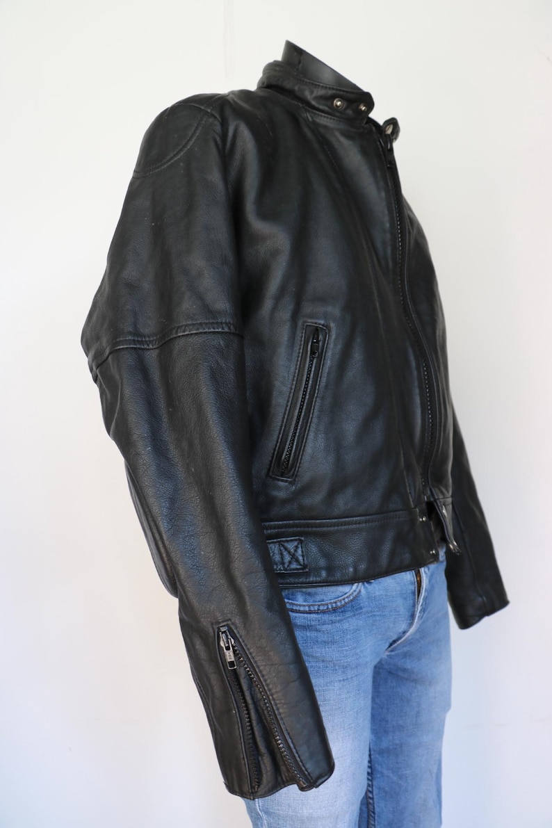 May include: Black leather motorcycle jacket with a stand-up collar, zippered pockets, and sleeve zippers. The jacket has a classic design with a fitted silhouette. The jacket is paired with blue jeans.