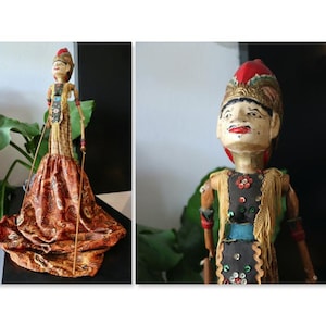 May include: A Javanese rod puppet featuring a hand-painted wooden head with a red headdress, a black bodice adorned with gold fringe and colourful sequins, and a long, flowing skirt with a brown and gold paisley pattern, manipulated by thin rods.
