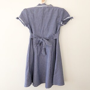 Puede incluir: Vestido de cuadros vichy azul y blanco con mangas cortas y cintura anudada en la espalda. El vestido est&aacute; colgado de una percha blanca sobre un fondo blanco. Cuello camisero y ribete blanco.