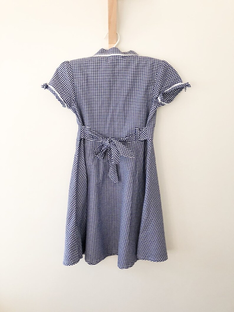 Puede incluir: Vestido de cuadros vichy azul y blanco con mangas cortas y cintura anudada en la espalda. El vestido est&aacute; colgado de una percha blanca sobre un fondo blanco. Cuello camisero y ribete blanco.