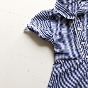 Puede incluir: Vestido de cuadros azul y blanco con cuello y ribete blancos. El vestido de manga corta tiene botones blancos en la parte delantera y est&aacute; hecho de una tela ligera. El vestido es probablemente vintage o retro.