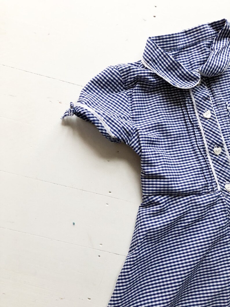 Puede incluir: Vestido de cuadros azul y blanco con cuello y ribete blancos. El vestido de manga corta tiene botones blancos en la parte delantera y est&aacute; hecho de una tela ligera. El vestido es probablemente vintage o retro.