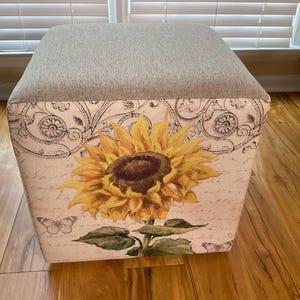 May include: Square ottoman with a light gray top and a decorative floral print on the sides. The print features a large yellow sunflower with green leaves and butterflies against a cream background.