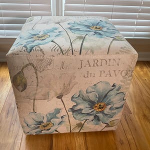 May include: Square ottoman with a floral print featuring large blue flowers with yellow centers on a cream background. The fabric has a vintage look with the words "JARDIN du PAVOT" printed on the side.