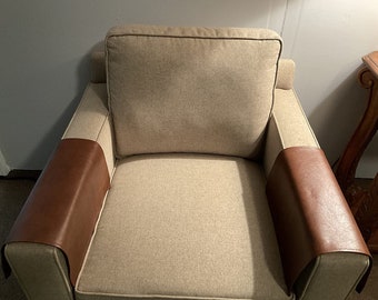 May include: A beige armchair with brown leather armrest covers. The chair has a tufted back and a thick, padded seat.