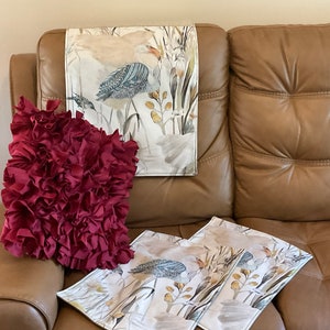 May include: A brown leather sofa with three throw pillows. Two of the pillows have a floral pattern with birds and the third pillow is a deep red color with a ruffled texture.
