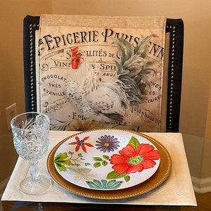 Puede incluir: Un plato de cena de cerámica blanca con un borde dorado presenta un diseño floral colorido con flores rojas, naranjas, verdes y moradas. El plato está colocado sobre un mantel individual blanco con un plato de servicio dorado debajo.