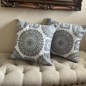 May include: Two square decorative pillows with a white background and a gray mandala pattern. The pillows are placed on a beige tufted bench. A decorative framed mirror hangs in the background.