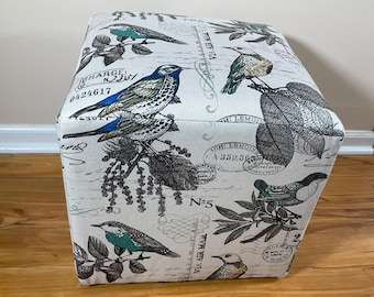 May include: Square ottoman with a cream-colored fabric featuring a bird and botanical print. The design includes blue, teal, and brown birds, along with leaves and vintage postage-style text. The ottoman is a cube shape.