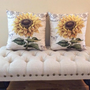 May include: Two square throw cushions with a white background and a sunflower design. The sunflowers are yellow with brown centres and green leaves. The cushions have a white background with a black and grey floral pattern.