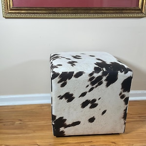 Könnte beinhalten: Ein quadratischer Hocker mit Kuhfellmuster. Der Hocker ist mit weißem Stoff mit unregelmäßigen schwarzen Flecken bezogen. Das Stück hat eine Würfelform und steht auf einem Holzboden.