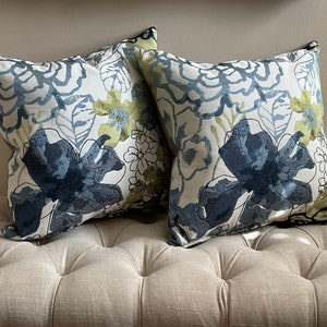 May include: Two decorative throw pillows with a floral pattern in shades of blue, green, and white. The pillows are on a light beige couch.