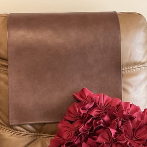 May include: A brown leather headrest cover for a chair or sofa. The cover is placed on top of a tan leather chair with a red ruffled pillow in front of it.