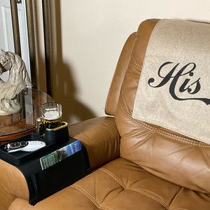 May include: A brown leather recliner with a beige armrest cover that says "His" in black script. A black armrest tray with a remote control, a magazine, and a glass of beer is on the armrest.
