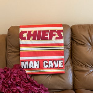Könnte beinhalten: Eine braune Ledersofa mit einer rot-weiß-goldenen Chiefs Man Cave Flagge, die über die Rückenlehne gelegt ist. Ein burgunderfarbenes, gerüschtes Kissen befindet sich auf der linken Seite des Sofas.