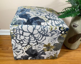 May include: A square ottoman upholstered in a fabric featuring a large-scale floral pattern. The design displays dark blue, light blue, and olive green flowers and leaves against a white background. The fabric has a visible woven texture.