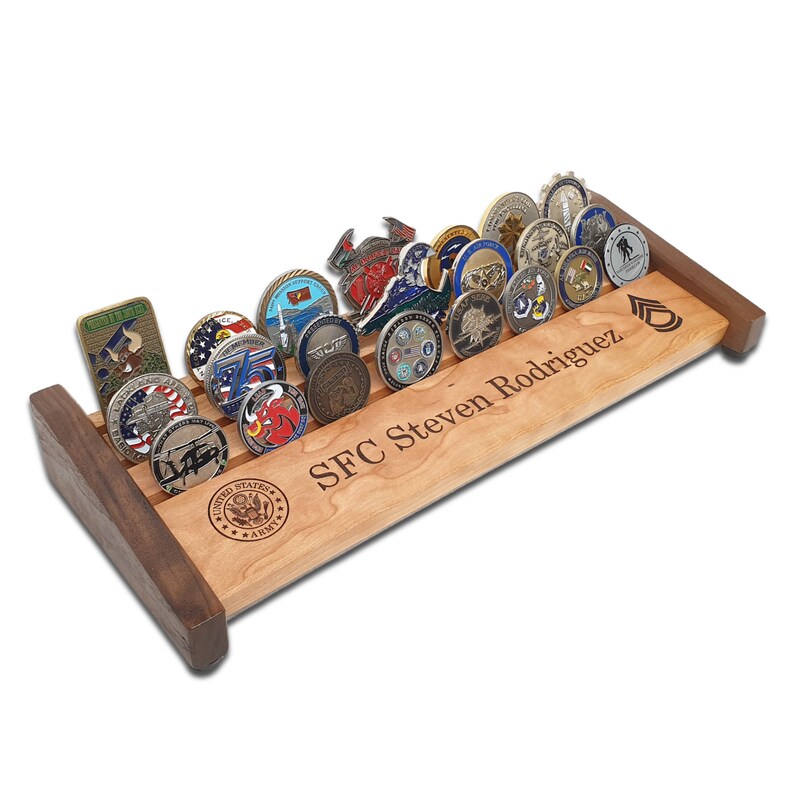 May include: A wooden coin display rack with a personalized inscription "SFC Steven Rodriguez" and a United States Army logo. The rack holds a collection of military challenge coins.
