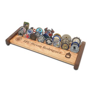 May include: A wooden coin display rack with a personalized inscription "SFC Steven Rodriguez". The rack is made of light brown wood with dark brown accents and features a row of military challenge coins.