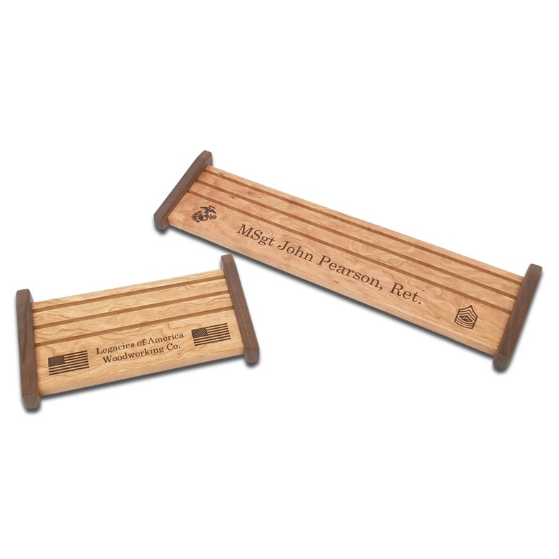 May include: Two wooden display stands with three slots for medals or coins. The top stand is engraved with "MSgt John Pearson, Ret." and features a US Marine Corps emblem and a military rank insignia. The bottom stand is engraved with "Legacies of America Woodworking Co." and features two American flags.