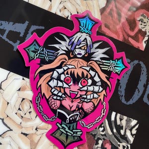 Puede incluir: Una pegatina rosa con un diseño en forma de cruz que presenta un personaje de dibujos animados con ojos rojos, cadenas y un segundo personaje encima. La pegatina tiene un fondo negro con otras ilustraciones.