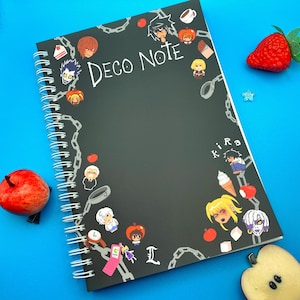 May include: Black spiral-bound notebook with the text "DECO NOTE" in white. The cover is decorated with cartoon characters, chains, and small illustrations. A red apple and a strawberry are on a blue surface.
