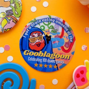 May include: A round button with the text "I got a brain eating amoeba at the Gooblagooon". The button features a cartoon character in a blue hooded robe and sunglasses, with a black cat. The button is on an orange background.