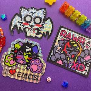 May include: A collection of holographic stickers featuring cartoon animals with an emo aesthetic. One sticker shows a bat-like creature, another says "I ❤️ EMOS", and a third says "RAWR XD". The stickers are on a purple background.
