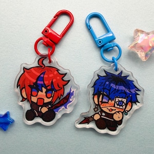 Op de afbeelding: Twee acryl sleutelhangers in anime-stijl. De ene sleutelhanger heeft een personage met rood haar en een rode sluiting, de andere heeft blauw haar en een blauwe sluiting. Beide hebben cartoon illustraties op een lichtblauwe achtergrond.