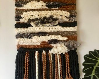 May include: A woven wall hanging with a brown, black, white, and gray color scheme. The hanging has a fringe at the bottom and is made with different textures of yarn.