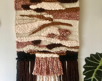 May include: A woven wall hanging with a brown, beige, and pink color scheme. The hanging features a variety of textures and patterns, including stripes, clouds, and swirls. The hanging has a fringe border.