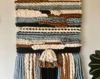 May include: A woven wall hanging with a geometric pattern in shades of blue, brown, white, and black. The hanging has a fringe border with tassels.
