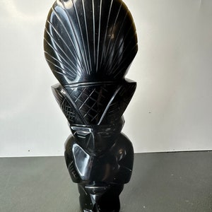May include: Black obsidian sculpture of a person with a large, carved shell headdress. The sculpture is detailed and has a smooth, polished finish.
