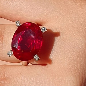 Puede incluir: Un anillo con una gran piedra de rubí de forma ovalada rodeada de cuatro diamantes más pequeños. El anillo está engastado en una banda de metal blanco.