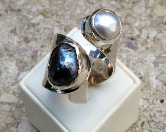 May include: A silver ring with a black and blue pearl and a white pearl. The ring is on a white display stand.