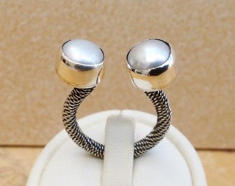 May include: A silver-toned ring featuring two large, round, white pearls. The ring's band has a woven, textured design. The pearls are set in silver bezels, creating a modern and elegant look. The ring is displayed on a white stand.