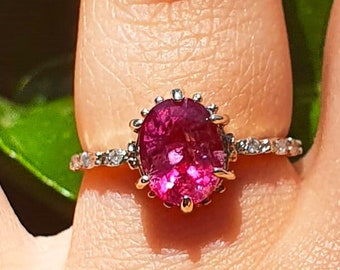 Puede incluir: Un anillo con una gran piedra preciosa rosa de forma ovalada rodeada de pequeñas piedras preciosas transparentes. El anillo está engastado en plata.