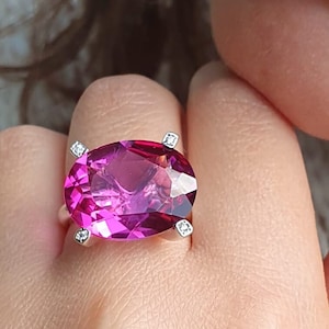 May include: A ring with a large oval-shaped pink gemstone surrounded by four smaller clear gemstones.