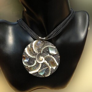 K&ouml;nnte beinhalten: Eine silberne Halskette mit einem gro&szlig;en, runden Anh&auml;nger, der ein schwarzes und irisierendes Abalone-Muschel-Design zeigt. Der Anh&auml;nger ist spiralf&ouml;rmig und &auml;hnelt einer Nautilus-Schale.