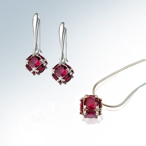 Puede incluir: Un conjunto de joyas de piedras preciosas de color rojo rubí que incluye un collar con colgante y pendientes a juego. El colgante y los pendientes presentan un diseño redondo y enjaulado con una piedra preciosa roja en el centro.