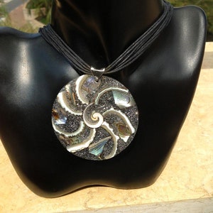 K&ouml;nnte beinhalten: Eine silberne Halskette mit einem gro&szlig;en, runden Anh&auml;nger, der ein Spiralmuster aus schimmerndem Abalone-Muschel und schwarzem Glitzer zeigt. Der Anh&auml;nger ist an einer schwarzen Schnurkette befestigt.