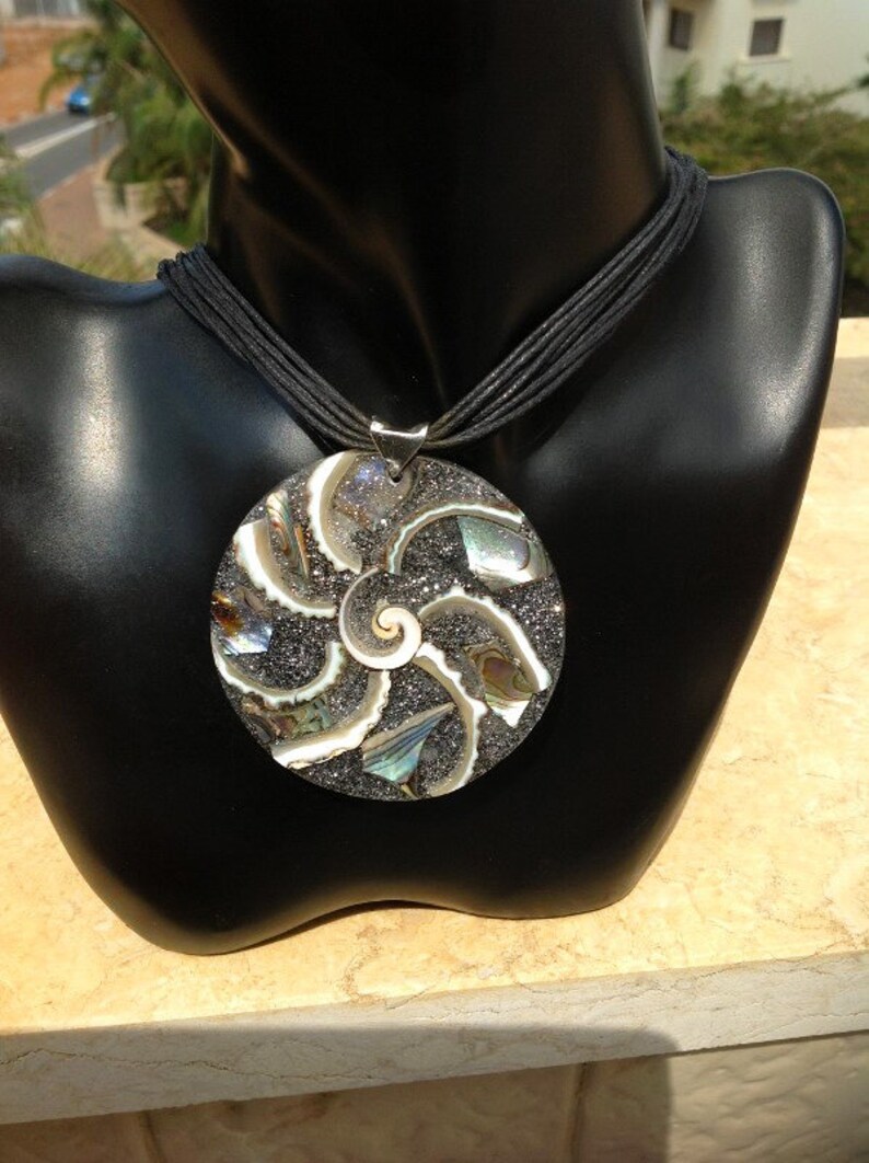 K&ouml;nnte beinhalten: Eine silberne Halskette mit einem gro&szlig;en, runden Anh&auml;nger, der ein Spiralmuster aus schimmerndem Abalone-Muschel und schwarzem Glitzer zeigt. Der Anh&auml;nger ist an einer schwarzen Schnurkette befestigt.