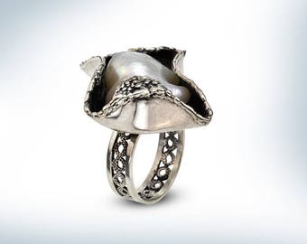 May include: A silver ring with a large, irregular pearl set in a delicate, ornate design. The ring band is also intricately detailed.