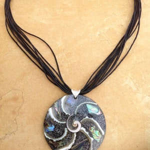 K&ouml;nnte beinhalten: Eine schwarze Schnurkette mit einem runden Anh&auml;nger, der ein Spiraldesign aus irisierendem Abalone-Muschel zeigt.